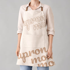 Stockka full length apron in beige cotton-polyester blend with cross straps and front pocket. Features 'apron mo' text graphic.