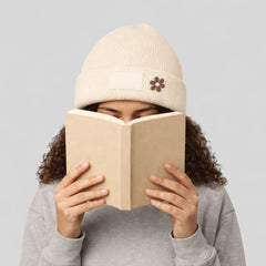 Stockka cream acrylic knit beanie with a small flower detail and a blank patch, worn by a person reading a book.