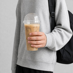 Stockka tall clear plastic cup with dome lid filled with iced coffee, held by a person wearing a grey sweatshirt.