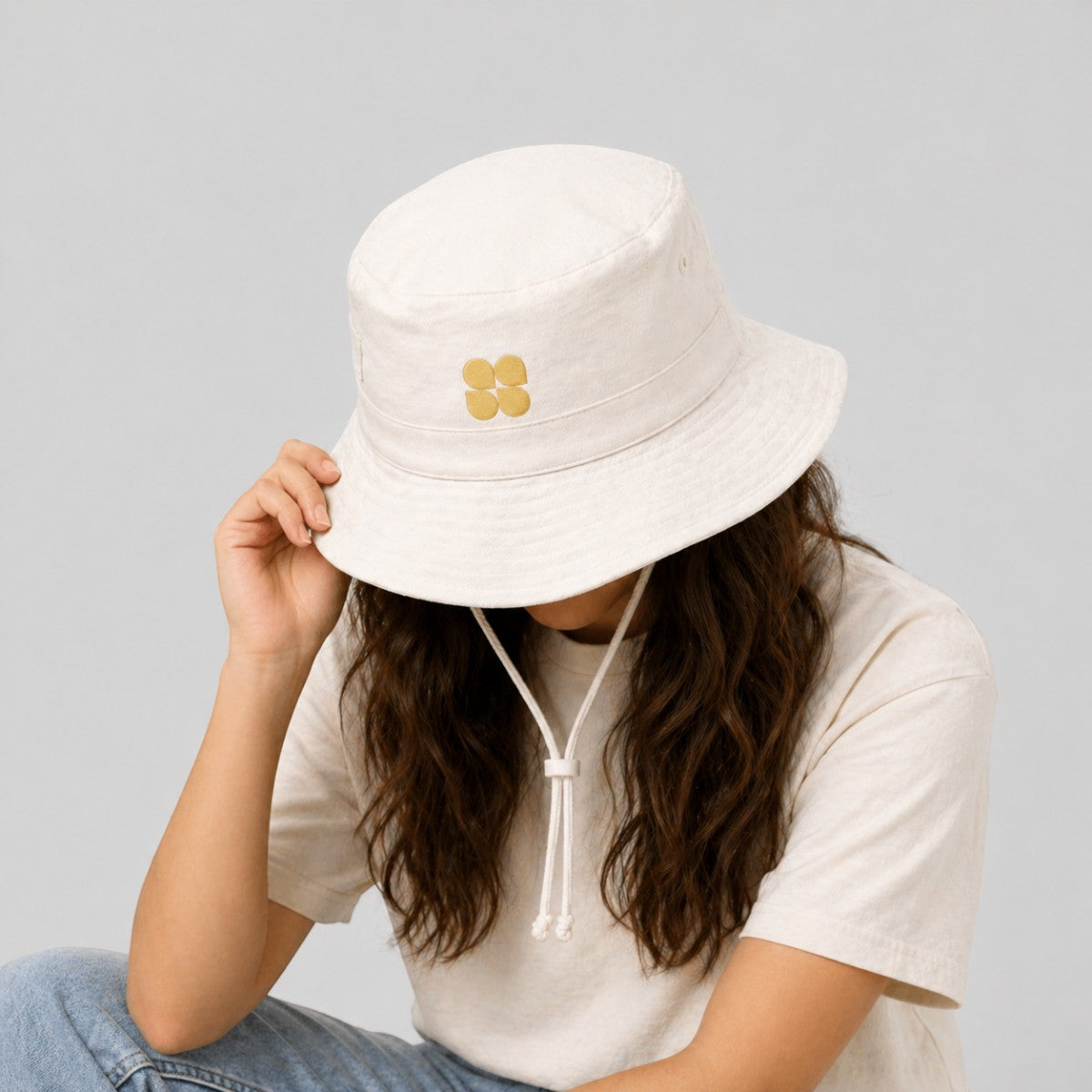 Stockka cream cotton blend bucket hat with gold four-petal flower logo and chin strap, ideal for beach or outdoor activities.
