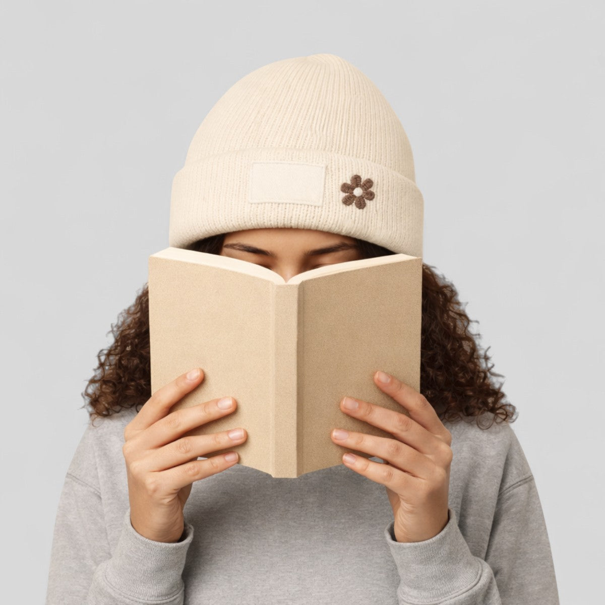 Stockka cream acrylic knit beanie with a small flower detail and a blank patch, worn by a person reading a book.