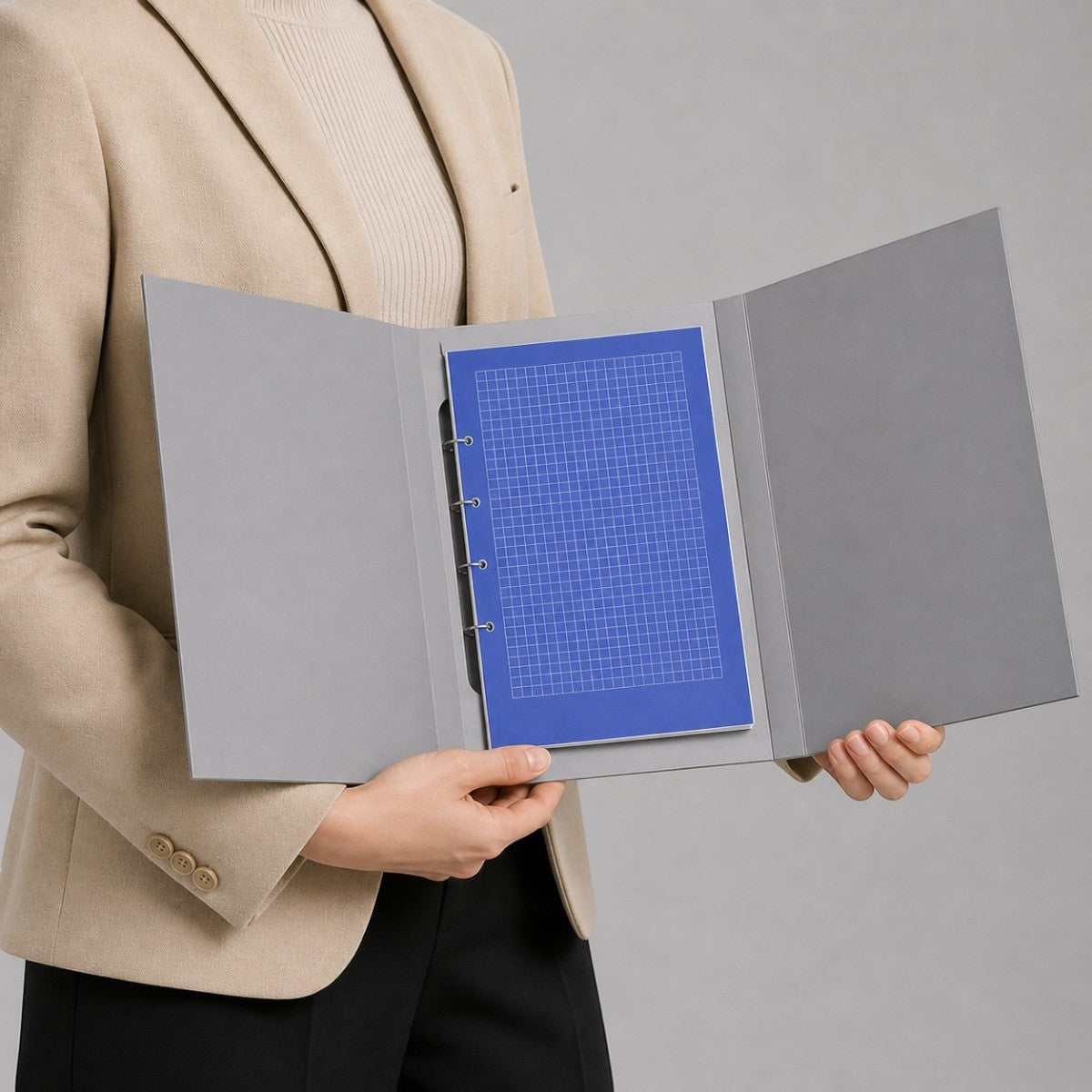 Stockka grey hard cover 3-fold binder with silver rings and blue grid paper insert, held by a person in a beige blazer.