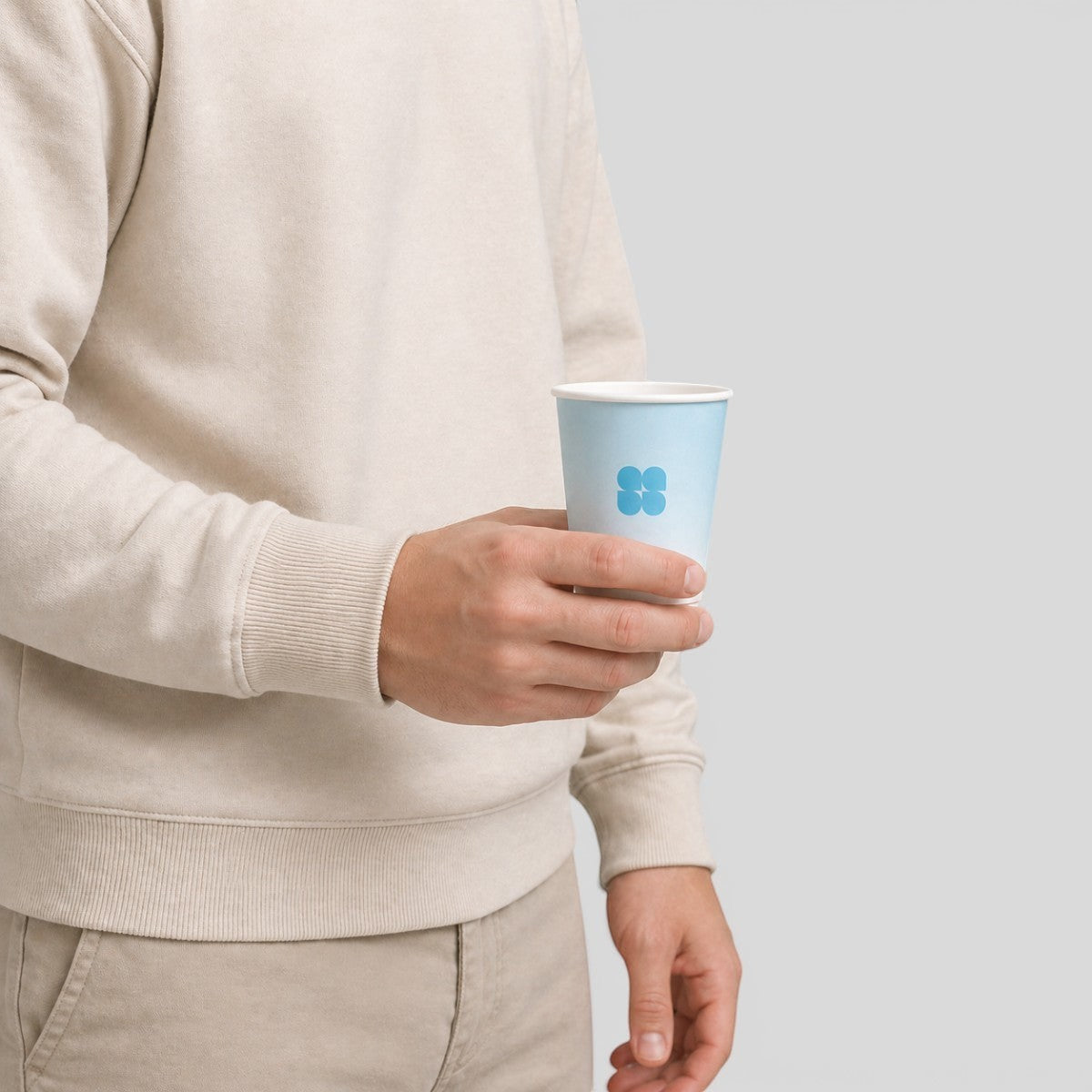 Stockka light blue paper cup with a blue logo, held by a person in a beige sweatshirt and tan pants.