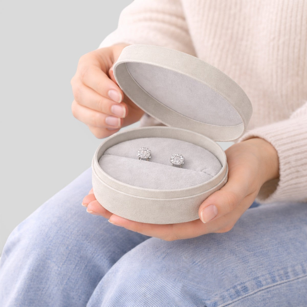 Stockka light grey round jewelry box with removable lid, holding diamond stud earrings. Made of rigid cardboard with matte finish.