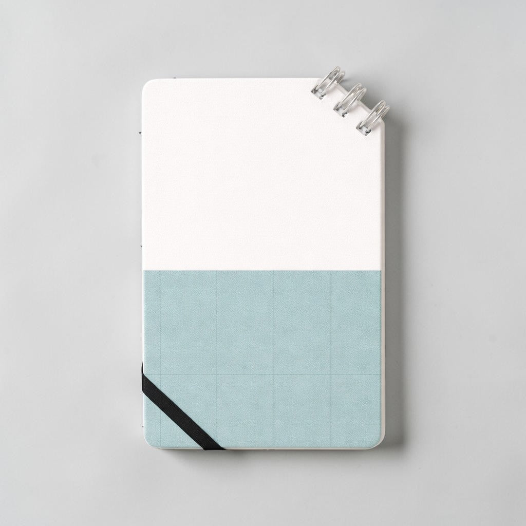 Stockka compact notebook with spiral binding at the top corner, elastic closure, and a two-tone design with a white top and blue grid bottom.