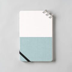 Stockka compact notebook with spiral binding at the top corner, elastic closure, and a two-tone design with a white top and blue grid bottom.